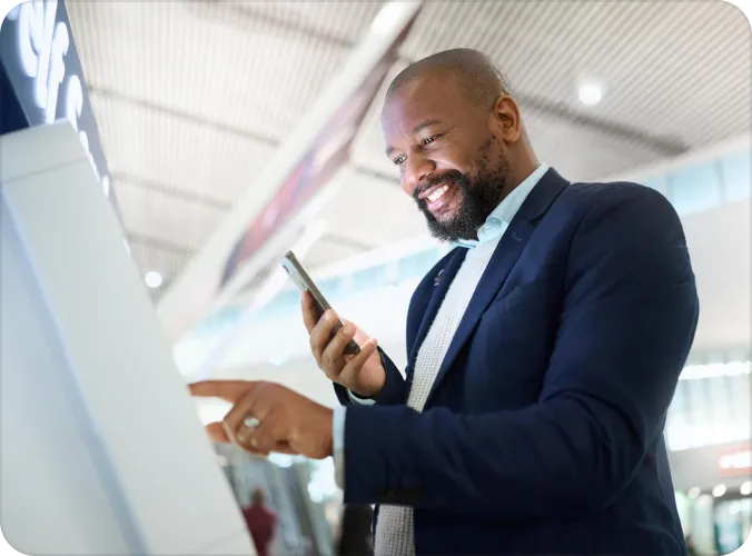 A smiling young man in a blazer hands cash to a bank teller across a counter at a financial institution, with a computer monitor visible in the background