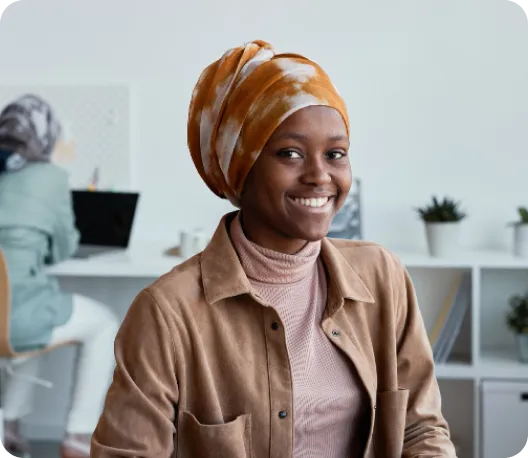 A young woman wearing an orange and white patterned head wrap and a tan jacket smiling warmly at the camera in a bright office environment, with colleagues visible in the background