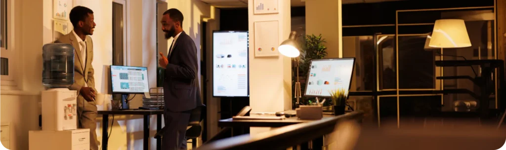 Two business professionals in suits engaged in a discussion in a modern office environment at night, with multiple screens displaying dashboards, data analytics charts, and deployed model visualizations in the background. A well-lit workstation with monitors showing graphs and reports is visible in the foreground