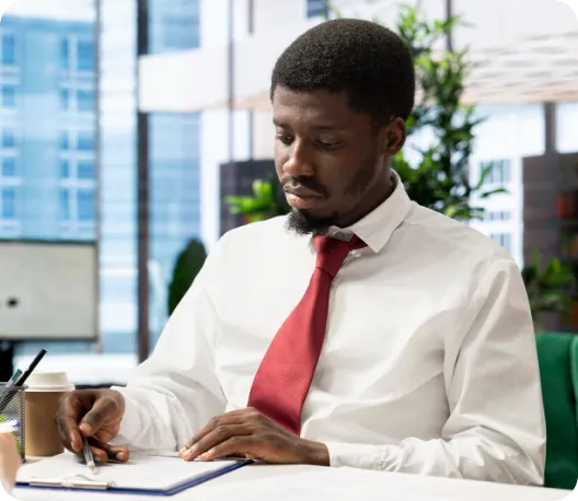A professional man in a white shirt and red tie seated at a desk, writing in a notebook with a coffee cup beside him and a city skyline visible through the window in the background