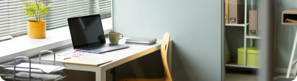 A neatly arranged office workstation featuring an open laptop, a coffee mug, a potted plant, stacked notebooks, and documents on a clean white desk beside a window with blinds, with a storage shelf and office partition visible in the background