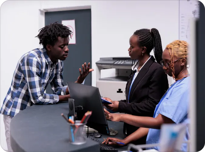 Three professionals — a man in a plaid shirt and two women, one in business attire and one in scrubs — collaborate around a desktop computer in an office setting, appearing to review information together