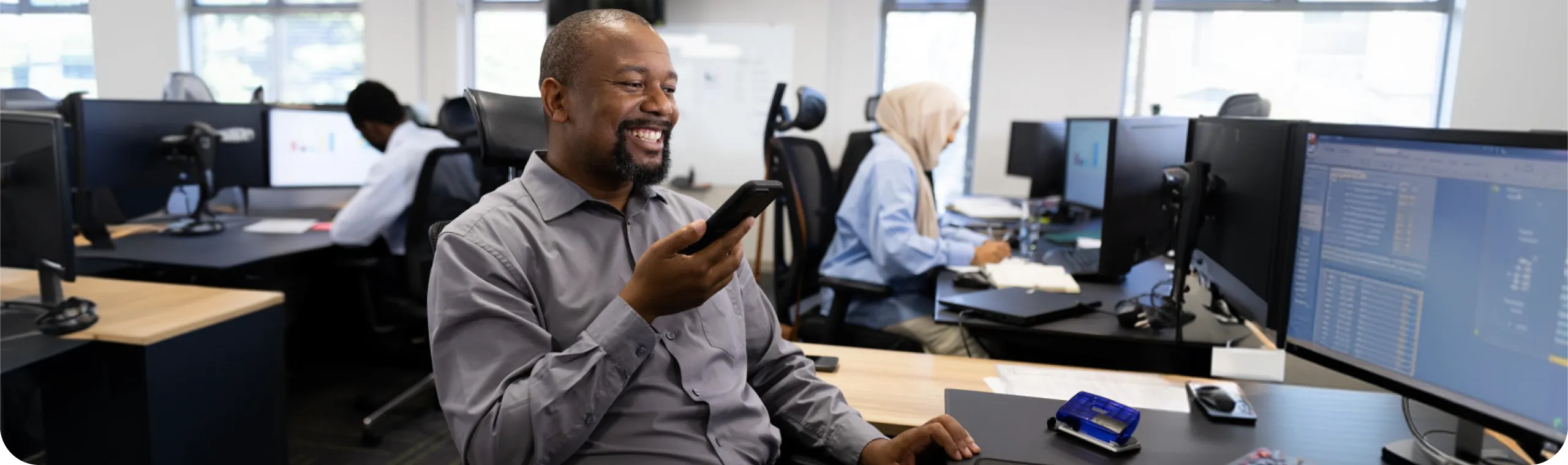 A smiling man in a grey shirt seated at his desk, holding a smartphone and laughing in a busy open-plan office, with colleagues working at computer workstations and multiple monitors displaying data dashboards visible in the background.