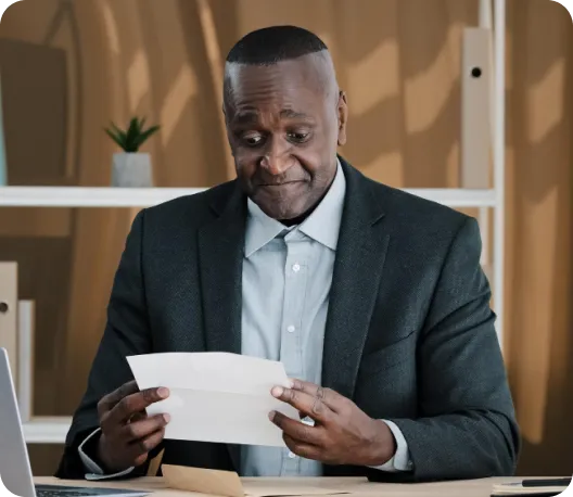 A middle-aged man in a dark blazer seated at a desk, carefully reading a document or letter with a thoughtful expression in a warmly lit office setting