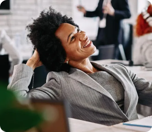 A woman in a grey blazer leaning back in her chair, smiling and looking upward confidently in a bright modern office environment
